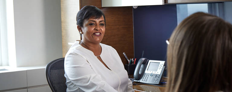 Lazard Woman Sitting at Desk Talking to Colleague Across from Her